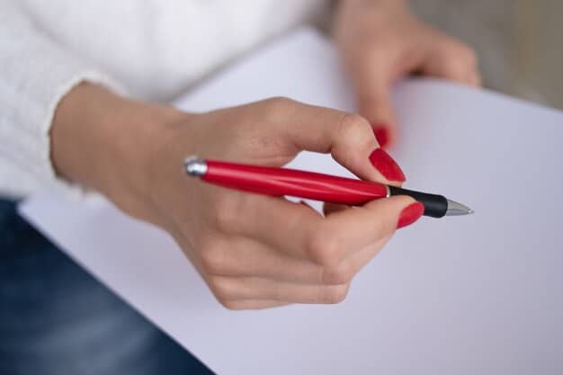 A person's hand holding a red pen, marking up a printed manuscript page with notes and corrections.
