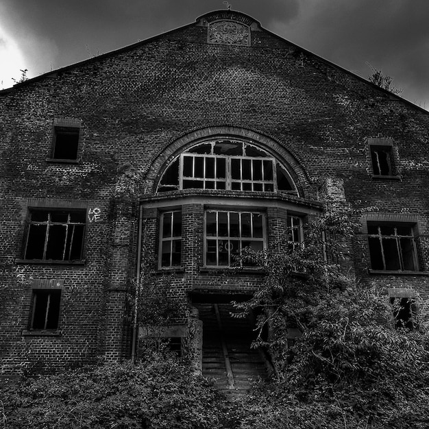 A black and white photograph of an old, gothic-style mansion with overgrown vines and darkened windows, conveying a sense of mystery and isolation. The sky is overcast, adding to the eerie atmosphere. This relates to Book Review: Unpacking Unreliable Narrators - 3 Novels That Will Keep You Guessing Until the End by visually representing the kind of setting where secrets and unreliable narrators thrive.